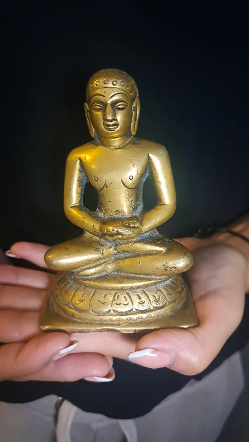 May include: A brass statue of a seated figure, possibly a deity or religious figure, in a meditative pose. The statue has a serene expression and intricate details. The figure is adorned with a headdress and sits on a lotus-shaped base. The statue is golden in color with a slightly aged patina, suggesting it may be antique or vintage. The overall style is traditional and possibly of Asian origin.