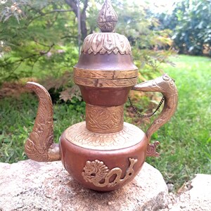 Könnte beinhalten: Eine dekorative Teekanne aus Messing und Kupfer mit einem Drachenmotiv. Die Teekanne hat einen Deckel mit einem Knauf und einen Ausguss in Form eines Vogelkopfes. Die Teekanne steht auf einem Stein.
