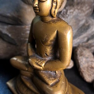 May include: A small, gold-colored brass statue of a seated Buddha. The statue is intricately detailed, with the Buddha's features and clothing clearly visible. The base of the statue is square and features decorative carvings. The statue is shown against a dark background.
