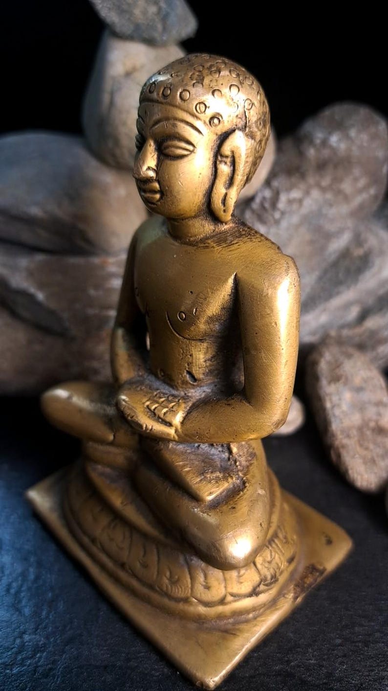 May include: A small, gold-colored brass statue of a seated Buddha. The statue is intricately detailed, with the Buddha's features and clothing clearly visible. The base of the statue is square and features decorative carvings. The statue is shown against a dark background.