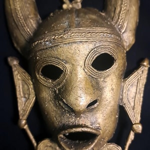 Puede incluir: Una máscara de metal de color dorado con un rostro humano, cuernos y detalles decorativos. La máscara tiene una boca grande abierta y ojos.