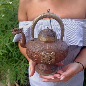 Könnte beinhalten: Eine Kupferteekanne mit einem Drachenausguss und aufwendigen floralen Designs. Die Teekanne hat einen silbernen Griff und eine silberne Kette, die am Deckel befestigt ist.