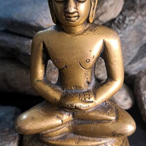 May include: A brass statue of a seated figure, possibly a deity or religious figure, in a meditative pose. The statue has a serene expression and intricate detailing on the head and base. The figure's torso is bare, and the hands are clasped in the lap. The statue is set against a dark background with stones.