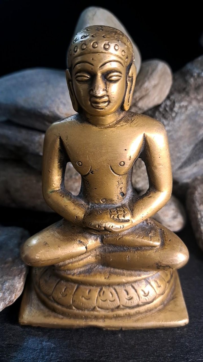 May include: A brass statue of a seated figure, possibly a deity or religious figure, in a meditative pose. The statue has a serene expression and intricate detailing on the head and base. The figure's torso is bare, and the hands are clasped in the lap. The statue is set against a dark background with stones.