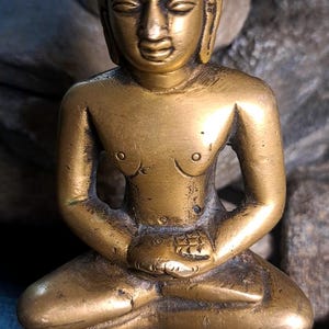 May include: A brass figurine of a seated figure, possibly a deity or spiritual leader, in a meditative pose. The figure has a serene expression and is adorned with intricate details on its head and clothing. The figurine is mounted on a square base with decorative carvings. The overall color is a rich, golden brass.  The statue's smooth surface shows signs of age and wear, adding to its antique appeal. This brass sculpture is a collectible item, ideal for home decor or spiritual practice.