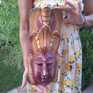 May include: A hand-carved wooden mask with a multi-coloured design featuring three snakes and a face. The mask is painted in shades of brown, yellow, and red.