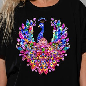 May include: Black t-shirt featuring a vibrant, jewel-toned peacock design. The peacock is rendered with a multitude of colorful gems in shades of blue, pink, purple, and green. The design is centered on the shirt, creating a striking visual.