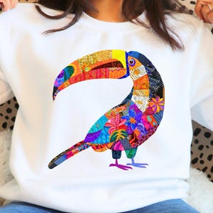 Op de afbeelding: Witte sweatshirt met een kleurrijk toekanontwerp. De toekan is samengesteld uit verschillende patronen in rood, oranje, geel, blauw en groen. De grote snavel en het lichaam van de vogel springen eruit tegen de witte achtergrond.