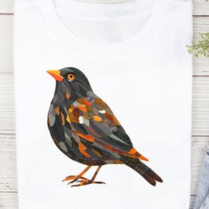 Op de afbeelding: Wit T-shirt met een gedetailleerde illustratie van een vogel. De vogel is afgebeeld met een mozaïekachtig ontwerp, met tinten zwart, grijs, oranje en bruin. De vogel heeft een oranje snavel en oog en staat op twee oranje poten.