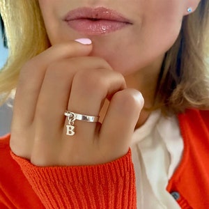 May include: A silver ring with a dangling letter 'B' charm. The ring is worn on a person's finger.