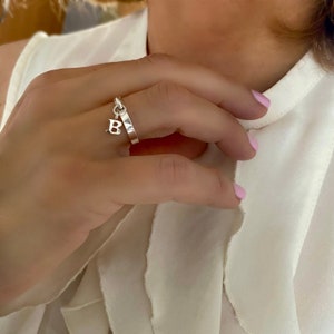 May include: A silver ring with a dangling letter 'B' charm. The ring is worn on a finger with pink nail polish.
