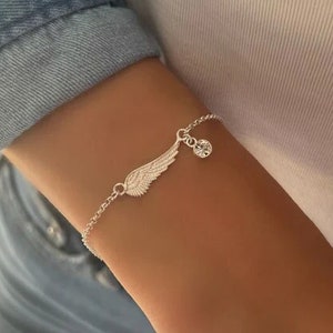 Puede incluir: Una pulsera de plata con un delicado encanto de ala de &aacute;ngel y un colgante de cristal transparente. La pulsera est&aacute; en la mu&ntilde;eca de una persona.