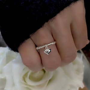 May include: A silver ring with a small heart charm dangling from it. The ring is worn on a finger.