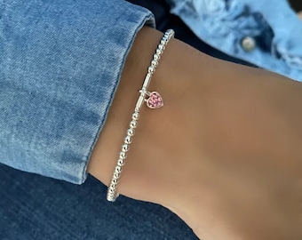 May include: A silver beaded bracelet with a pink heart charm. The bracelet is on a person's wrist.