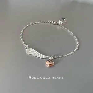 May include: A silver chain bracelet with a silver wing charm and a rose gold heart charm. The bracelet has an adjustable clasp. "ROSE GOLD HEART" is written below the bracelet.
