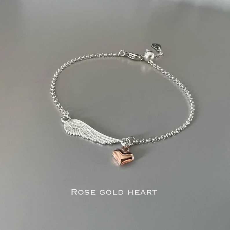 May include: A silver chain bracelet with a silver wing charm and a rose gold heart charm. The bracelet has an adjustable clasp. "ROSE GOLD HEART" is written below the bracelet.
