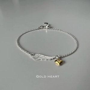 May include: Silver chain bracelet with a silver wing charm and a gold heart charm. The bracelet has a lobster clasp closure. The text "GOLD HEART" is printed below the bracelet.