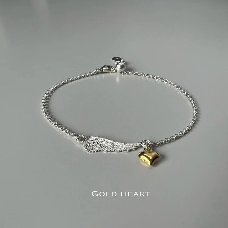 May include: Silver chain bracelet with a silver wing charm and a gold heart charm. The bracelet has a lobster clasp closure. The text "GOLD HEART" is printed below the bracelet.