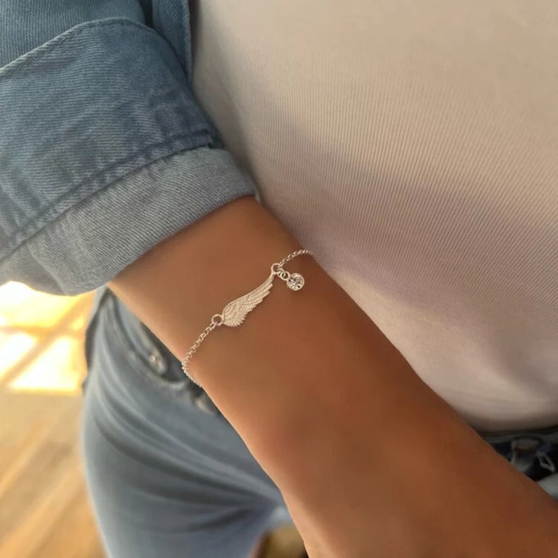 Puede incluir: Una pulsera de plata con un delicado encanto de ala de &aacute;ngel y un colgante de cristal transparente. La pulsera est&aacute; en la mu&ntilde;eca de una persona.
