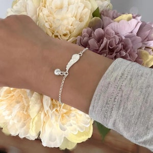 Puede incluir: Una pulsera de plata con un delicado encanto de ala de &aacute;ngel y una cuenta de cristal transparente. La pulsera est&aacute; en la mu&ntilde;eca de una persona.