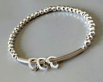May include: Silver beaded bracelet with three heart charms. The bracelet features small, round beads and a curved bar with three heart-shaped pendants. The bracelet is made of a shiny, metallic material.