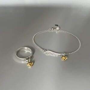 May include: A silver ring and bracelet set with a gold heart charm and a silver wing charm. The ring and bracelet are both delicate and dainty.