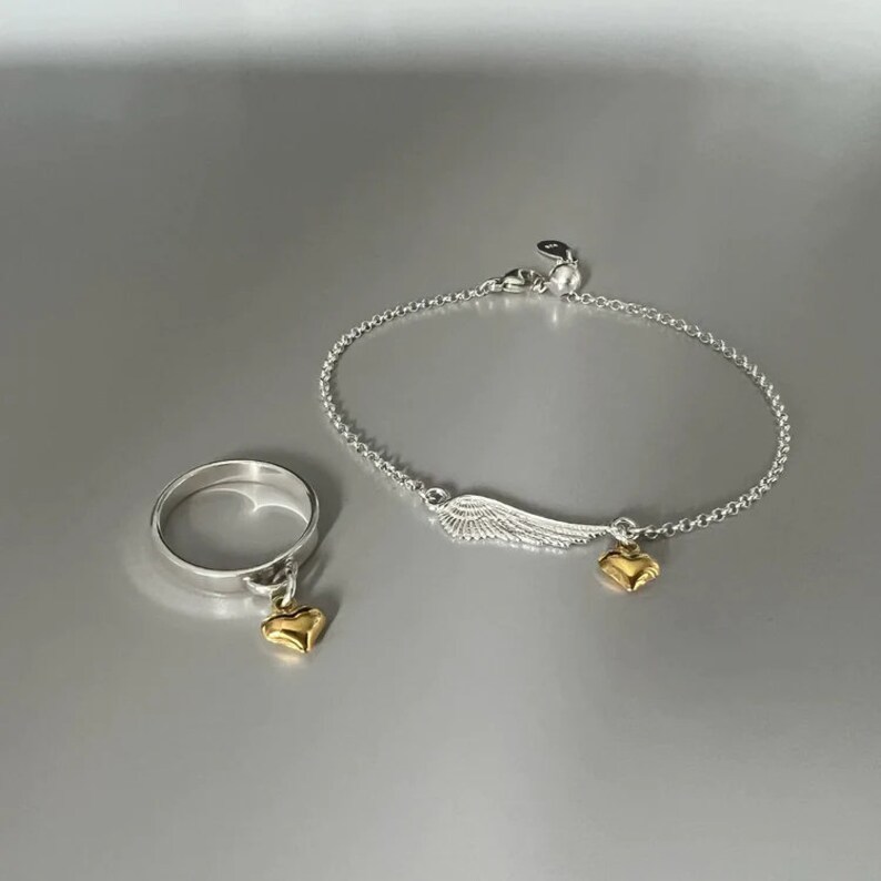 May include: A silver ring and bracelet set with a gold heart charm and a silver wing charm. The ring and bracelet are both delicate and dainty.
