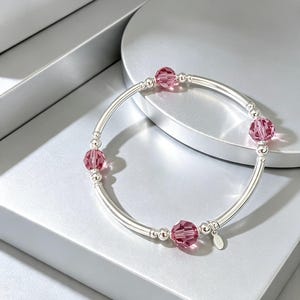 May include: A stretch bracelet featuring smooth silver-toned cylindrical beads and small round silver-toned spacer beads. The design is accented with four faceted, translucent pink crystal beads and a small, oval-shaped silver-toned metal charm.