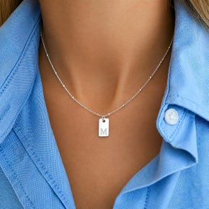 May include: A silver necklace featuring a delicate chain with small evenly spaced beads and a rectangular pendant. The pendant is engraved with the capital letter M in a serif font. The necklace is worn against a light blue collared shirt.