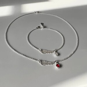 Puede incluir: Dos collares de plata con cadenas delicadas y colgantes de alas. El collar m&aacute;s peque&ntilde;o tiene un dije de cristal transparente, mientras que el collar m&aacute;s grande tiene un dije de piedra preciosa roja.