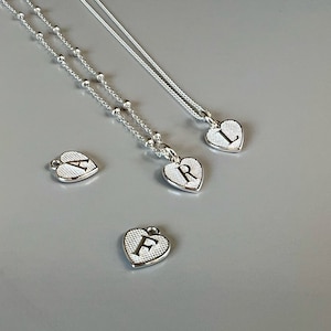 May include: Three silver necklaces with heart-shaped pendants featuring the letters A, R, and L. The necklaces are on a gray background.