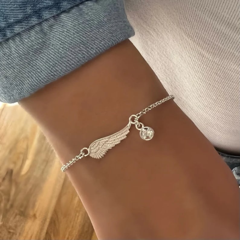 Puede incluir: Una pulsera de plata con una cadena delicada y un dije en forma de ala de &aacute;ngel con un acento de cristal transparente.