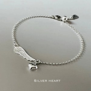 May include: A silver bracelet with a delicate chain and a charm featuring an angel wing and a small heart. The charm hangs from the chain and is a simple, elegant design.