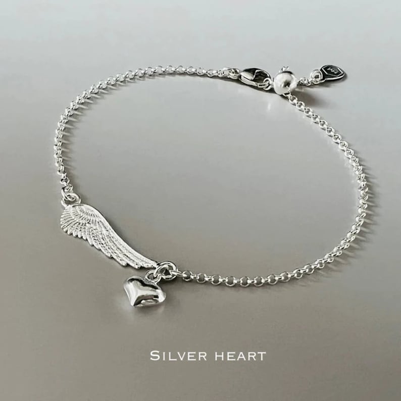 May include: A silver bracelet with a delicate chain and a charm featuring an angel wing and a small heart. The charm hangs from the chain and is a simple, elegant design.