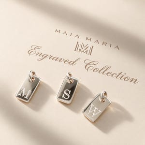 May include: Three rectangular gold-toned metal charms, each featuring a single engraved letter M, S, or W. Each charm includes a small circular jump ring at the top. The text MAIA MARIA Engraved Collection is printed above the charms.