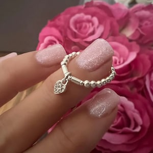 May include: A silver ring with a small heart charm. The heart charm is decorated with small clear crystals. The ring is on a finger with pink glitter nail polish.