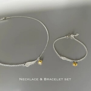 May include: A silver necklace and bracelet set with gold heart charms and silver angel wing charms.  The necklace and bracelet are both on a gray background.  "NECKLACE & BRACELET SET" is written in black text below the jewelry.