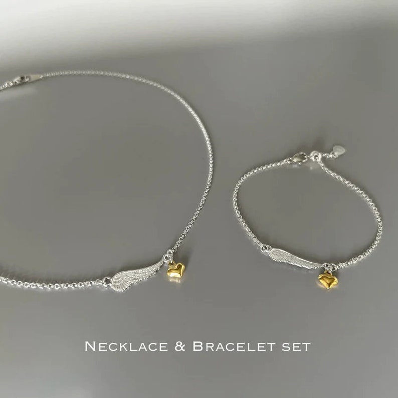 May include: A silver necklace and bracelet set with gold heart charms and silver angel wing charms.  The necklace and bracelet are both on a gray background.  "NECKLACE & BRACELET SET" is written in black text below the jewelry.