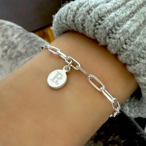 May include: A silver link bracelet with a circular charm. The charm features the letter "R" in a serif font. The bracelet is made of shiny, polished metal and is worn on a wrist.