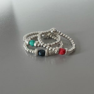 May include: Three silver beaded rings with colourful beads. The rings are stacked on top of each other. The beads are red, green, and black.