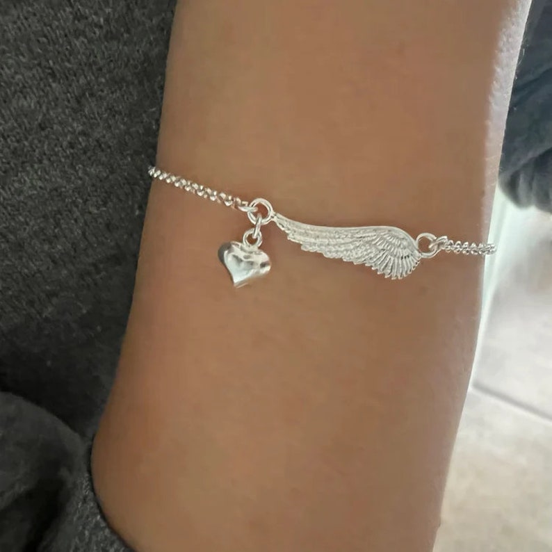 May include: A silver bracelet with a heart charm and a wing charm. The bracelet is on a person's wrist.