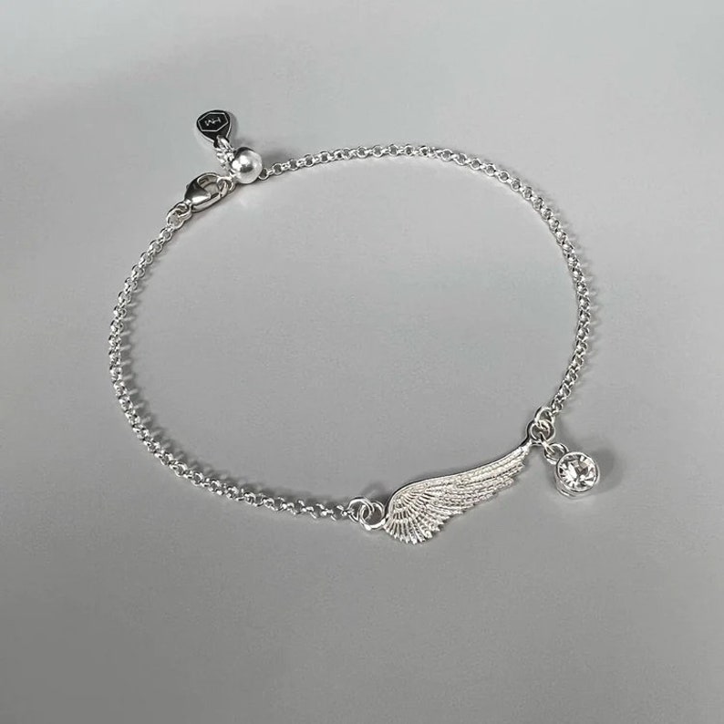 Puede incluir: Un brazalete de cadena de plata con un delicado encanto de ala y un colgante de cristal transparente.