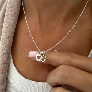 May include: A silver necklace featuring a delicate chain with small evenly spaced beads and two small circular charms. One charm displays a cutout heart shape, while the other charm features the capital letter G in a classic serif font.