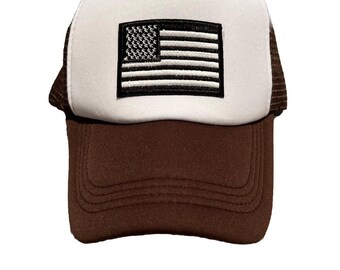 Op de afbeelding: Een truckerpet met een bruine klep en een wit voorpaneel. In het midden van het witte paneel bevindt zich een rechthoekige patch met een zwart-witte Amerikaanse vlag. De zijkanten en de achterkant zijn gemaakt van bruin gaas.