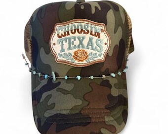 Op de afbeelding: Een camouflage truckerpet met een centrale patch met de tekst "CHOOSIN' TEXAS" in bruin en lichtblauw borduurwerk, geaccentueerd door een kleine oranje roos. Een fijn kettinkje met kleine lichtblauwe stenen kralen loopt over de voorkant.