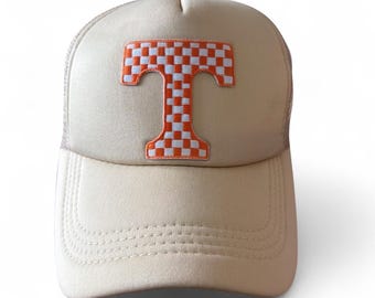 Op de afbeelding: Een beige truckerpet met een opvallende geborduurde letter T op het voorpaneel. De letter T is gevuld met een wit en oranje geblokt patroon. De pet heeft een gestructureerde voorkant, een gebogen klep met stiksels en een achterkant van mesh.