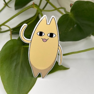 Op de afbeelding: Een gele cartoon-katten sticker met een vriendelijke uitdrukking, zwaaiend met zijn rechterpoot. De sticker is op een witte achtergrond met groene bladeren.