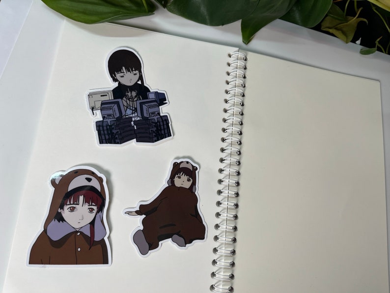 May include: Three anime-style stickers on a white page. The stickers feature a girl with short black hair wearing a white shirt and a black jacket, a girl with short brown hair wearing a brown bear costume, and a girl with short brown hair wearing a brown bear costume.