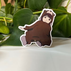 May include: A brown bear costume with a hood and a girl's face peeking out from the hood. The girl has brown hair and brown eyes. The costume is sitting on a white surface.