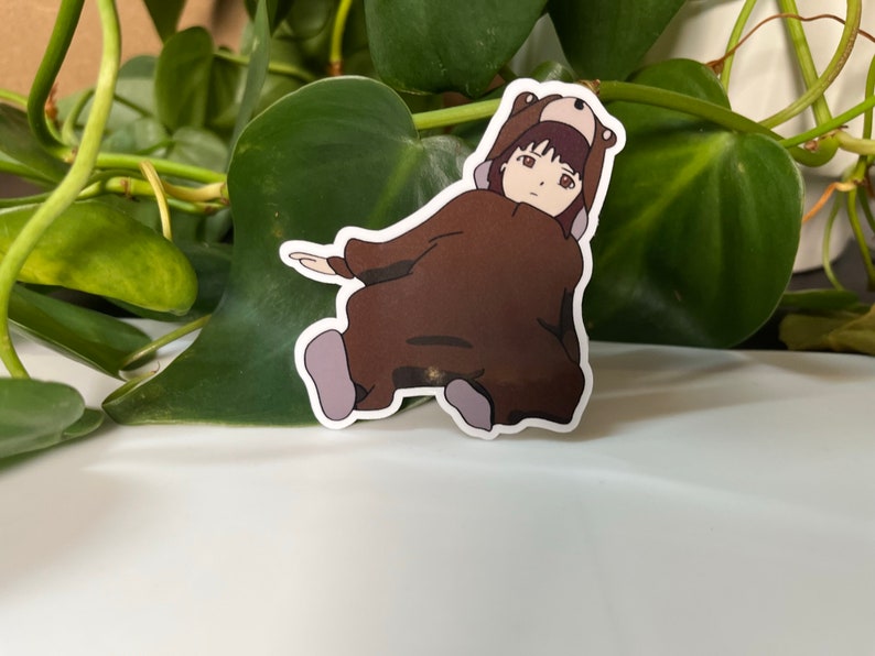 May include: A brown bear costume with a hood and a girl's face peeking out from the hood. The girl has brown hair and brown eyes. The costume is sitting on a white surface.